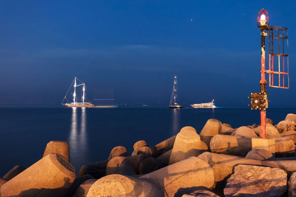 Night harbor with three sailboats on calm water; rocky breakwater in foreground and a red rusted beacon tower on the right.