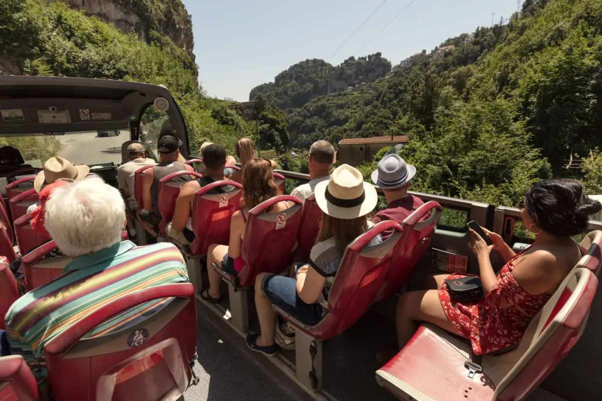 Open-top tour bus full of passengers wearing hats traveling along a hillside road past terraced houses.
