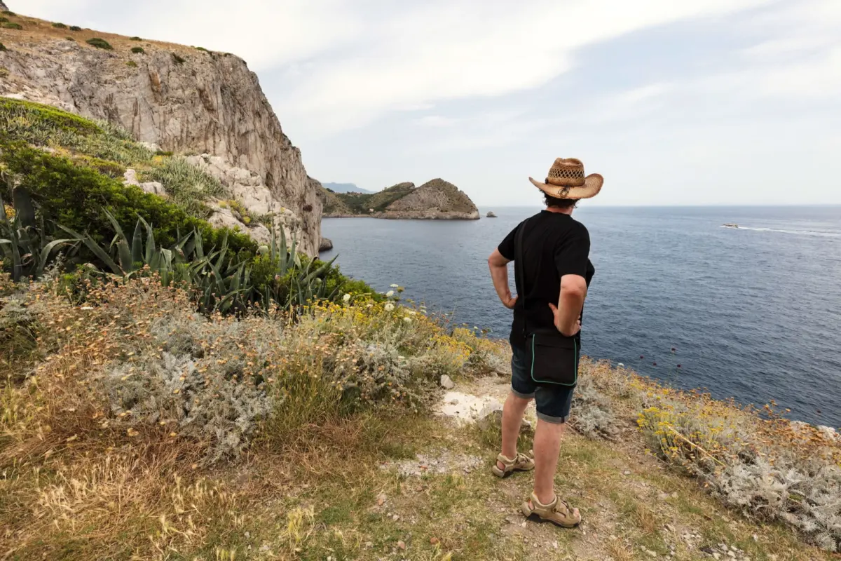 Adult man in straw hat, black shirt, and shorts stands with hands on hips on a coastal cliff edge overlooking the sea.