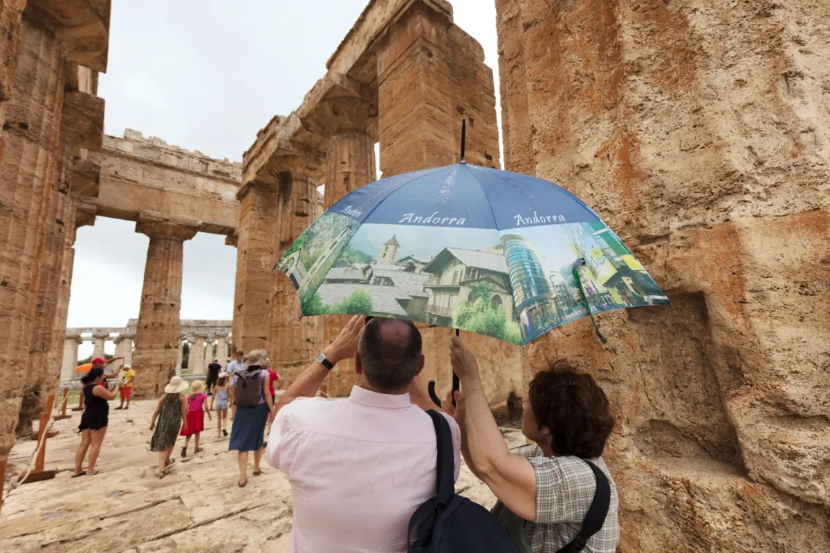 Tourists walk through ancient stone amphitheater ruins as a person holds a blue umbrella showing Andorra scenes.