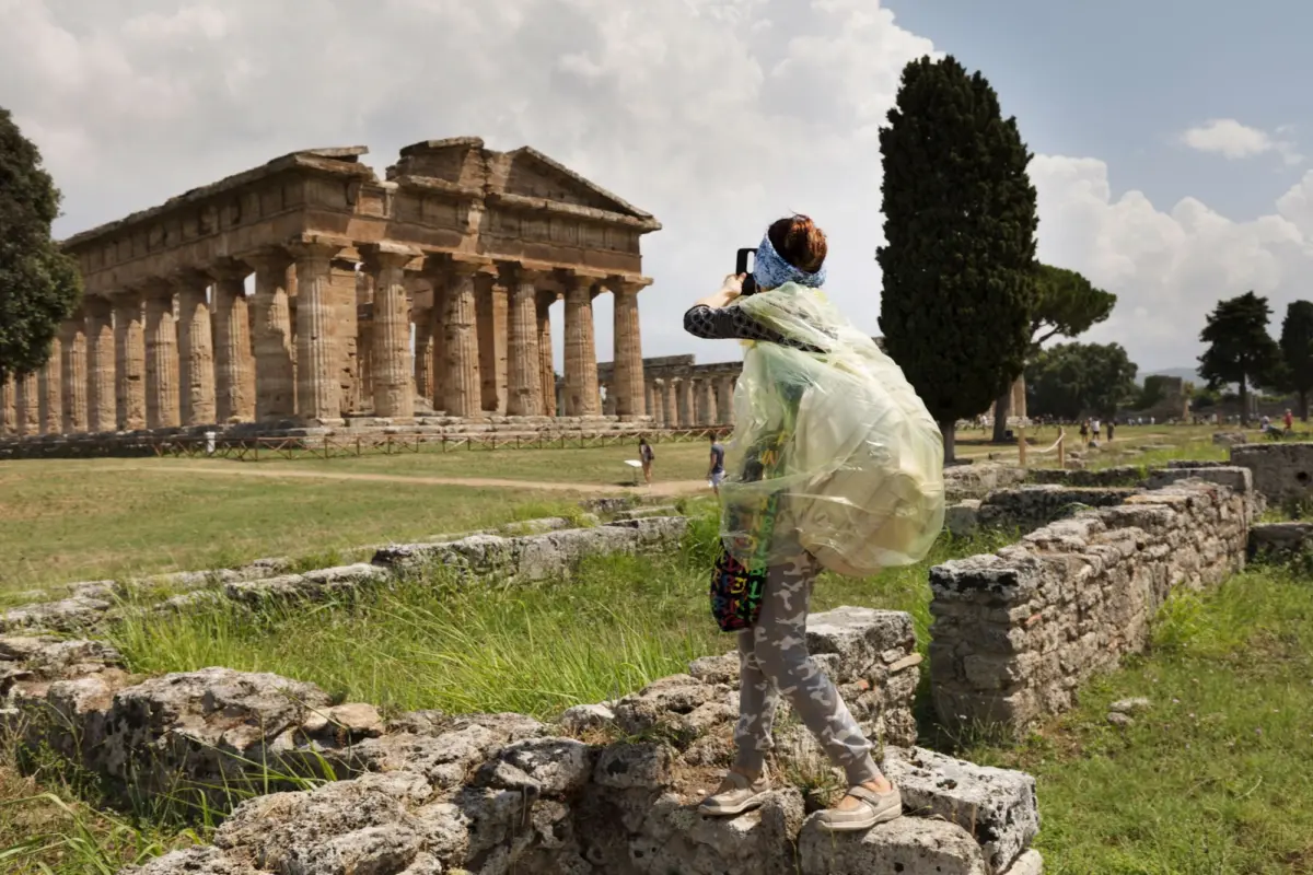 Person wearing a translucent rain poncho photographs ancient Greek temple ruins with a smartphone.