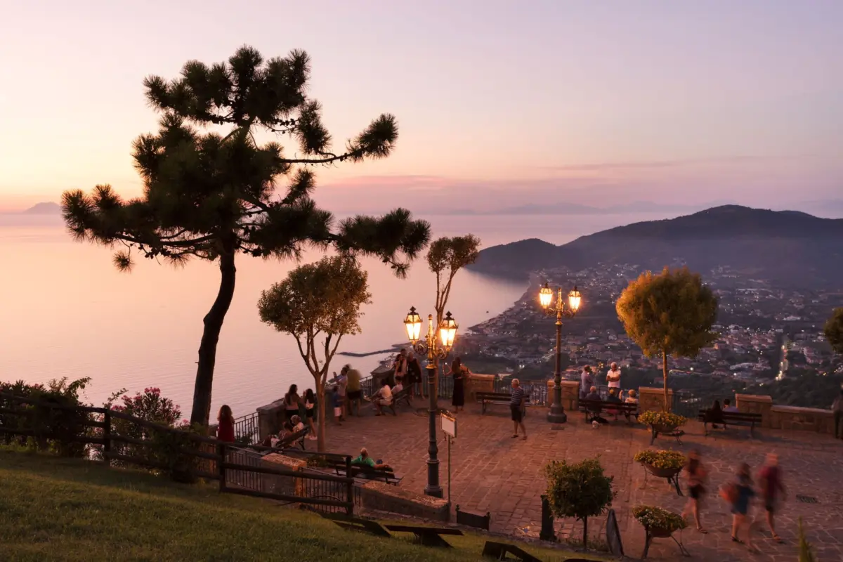 Silhouetted pine tree at a seaside overlook at sunset; lantern-lit terrace with visitors overlooking a pink-hued coastline and distant hills.