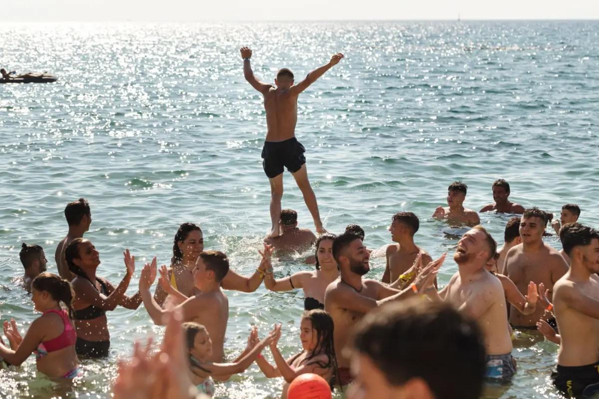 Person lifted on others' shoulders in the ocean, arms raised, amid cheering beachgoers.
