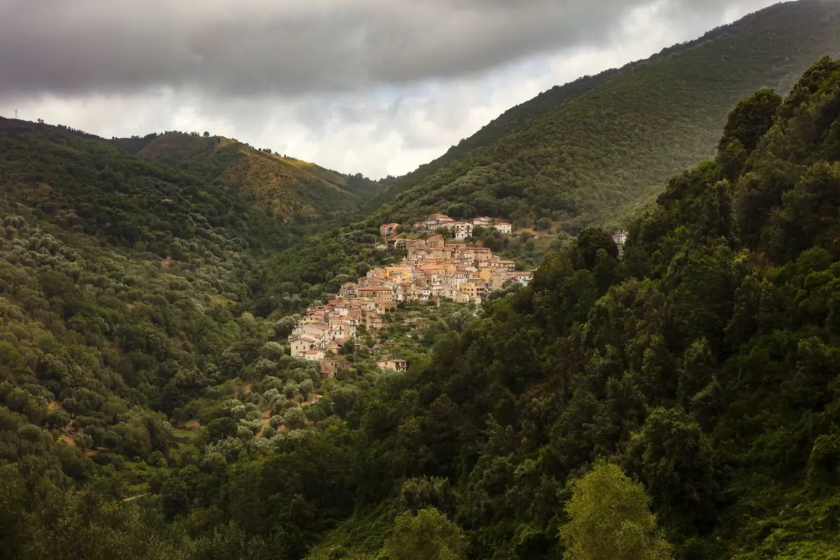 Hillside village of beige houses with red roofs nestled among green mountains and dense forests under a cloudy sky.