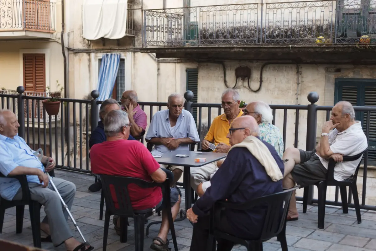 Group of elderly people seated around a table, playing cards on an outdoor terrace with ornate metal railing.
