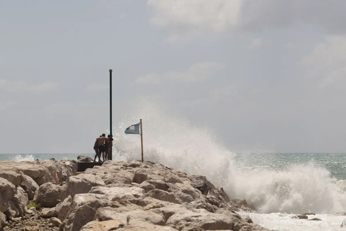 Group of people on a rocky jetty by the sea push a tall green pole as waves crash.