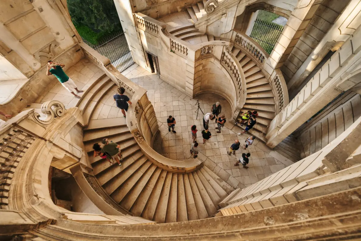 Tourists explore a grand circular stone staircase inside a historic palace.
