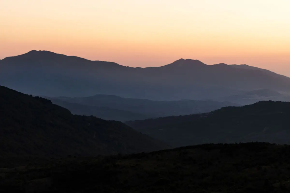 Dark mountain range at sunset with orange and pale yellow sky and hazy distant ridges.