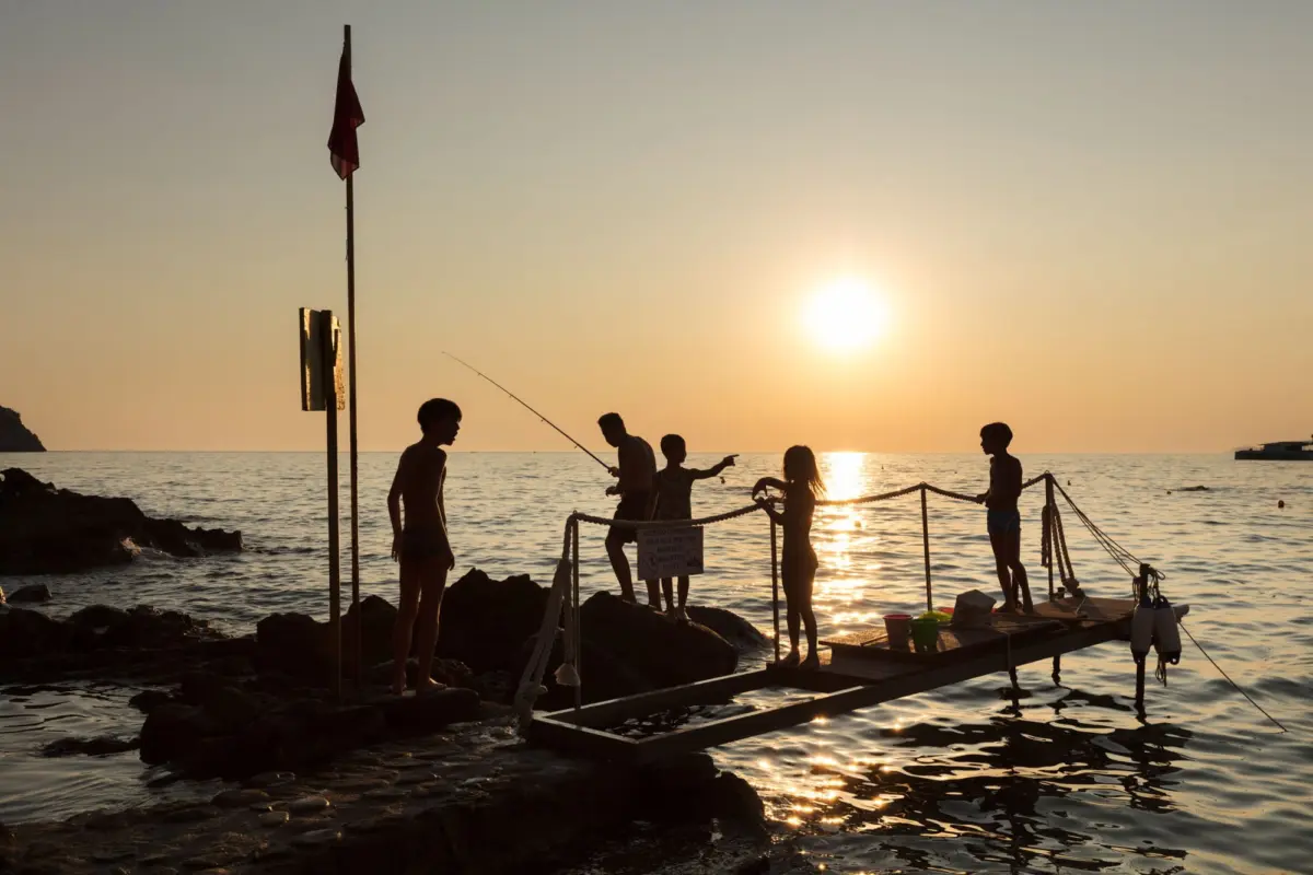 Silhouetted group of children fishing along a rope-lined rocky jetty at sunset.