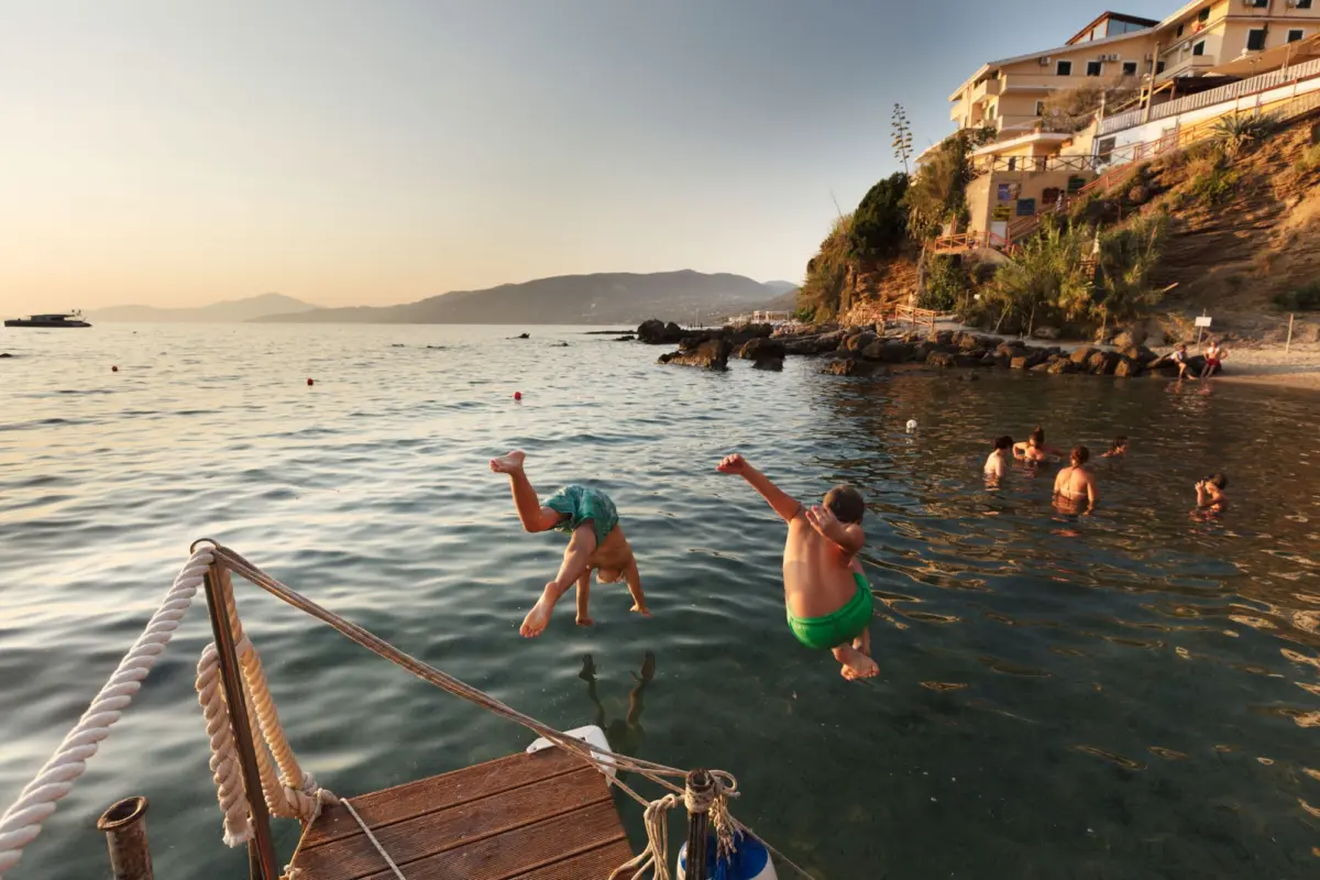 Children jump off a wooden dock into the sea along a rocky coastline with hillside buildings at sunset.