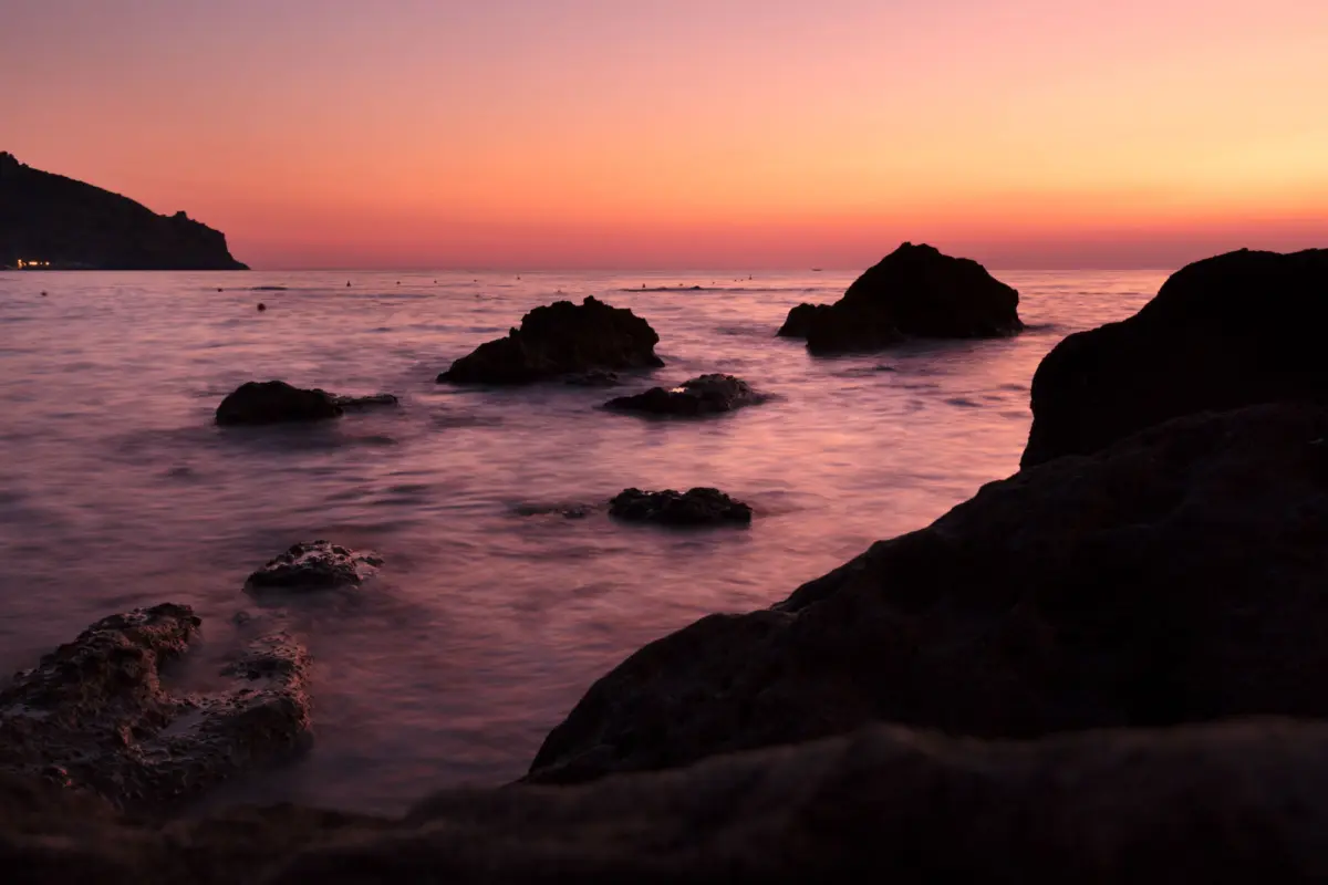 Silhouetted rocky shoreline at sunset with foreground rocks and pink-orange sky.