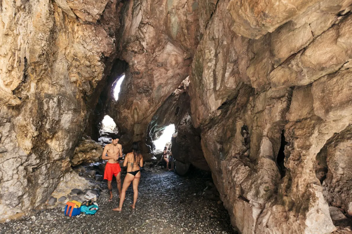 Group of people in swimwear inside a rugged sea cave, exploring near the light-filled opening.