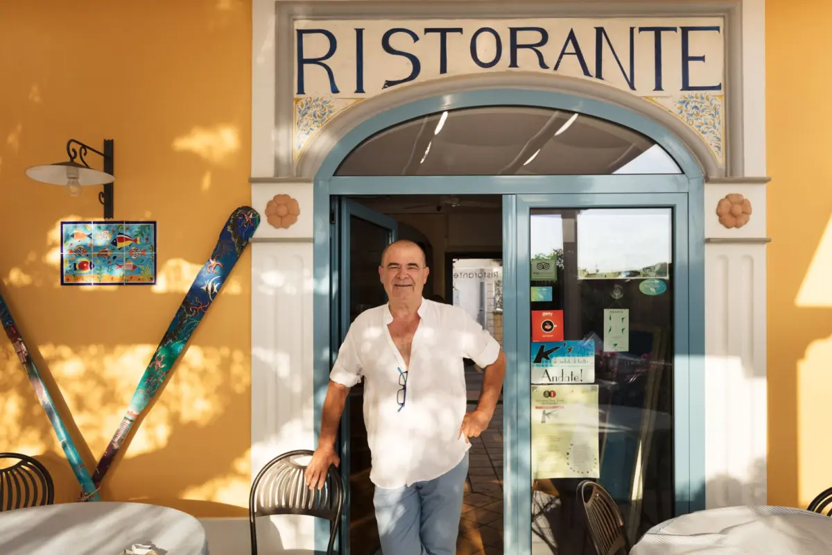 Smiling man in a white shirt stands in the teal doorway of a yellow Ristorante, with outdoor seating.