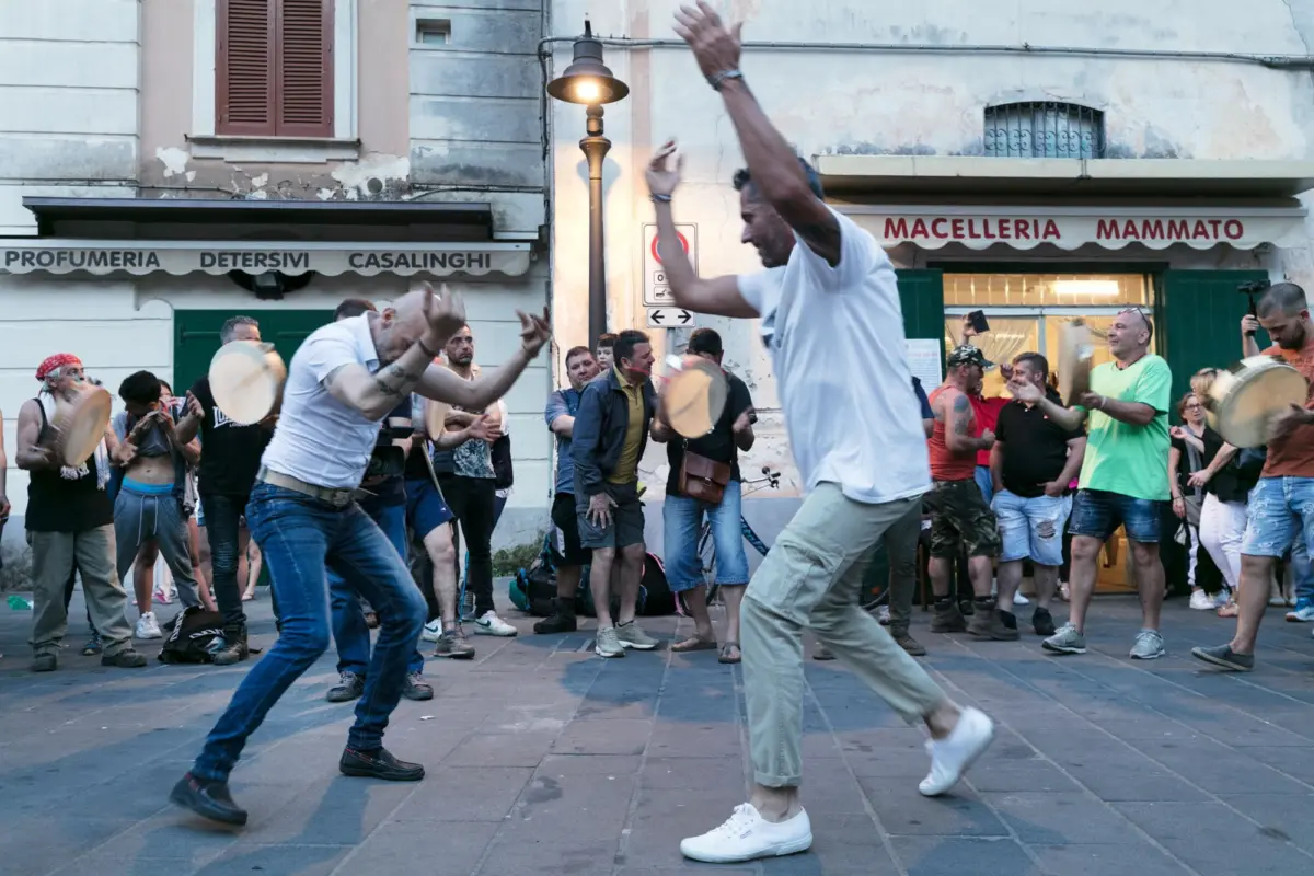 Dancer in white shirt raises arms amid crowd on busy Italian street; storefronts and the Macelleria Mammato sign visible.
