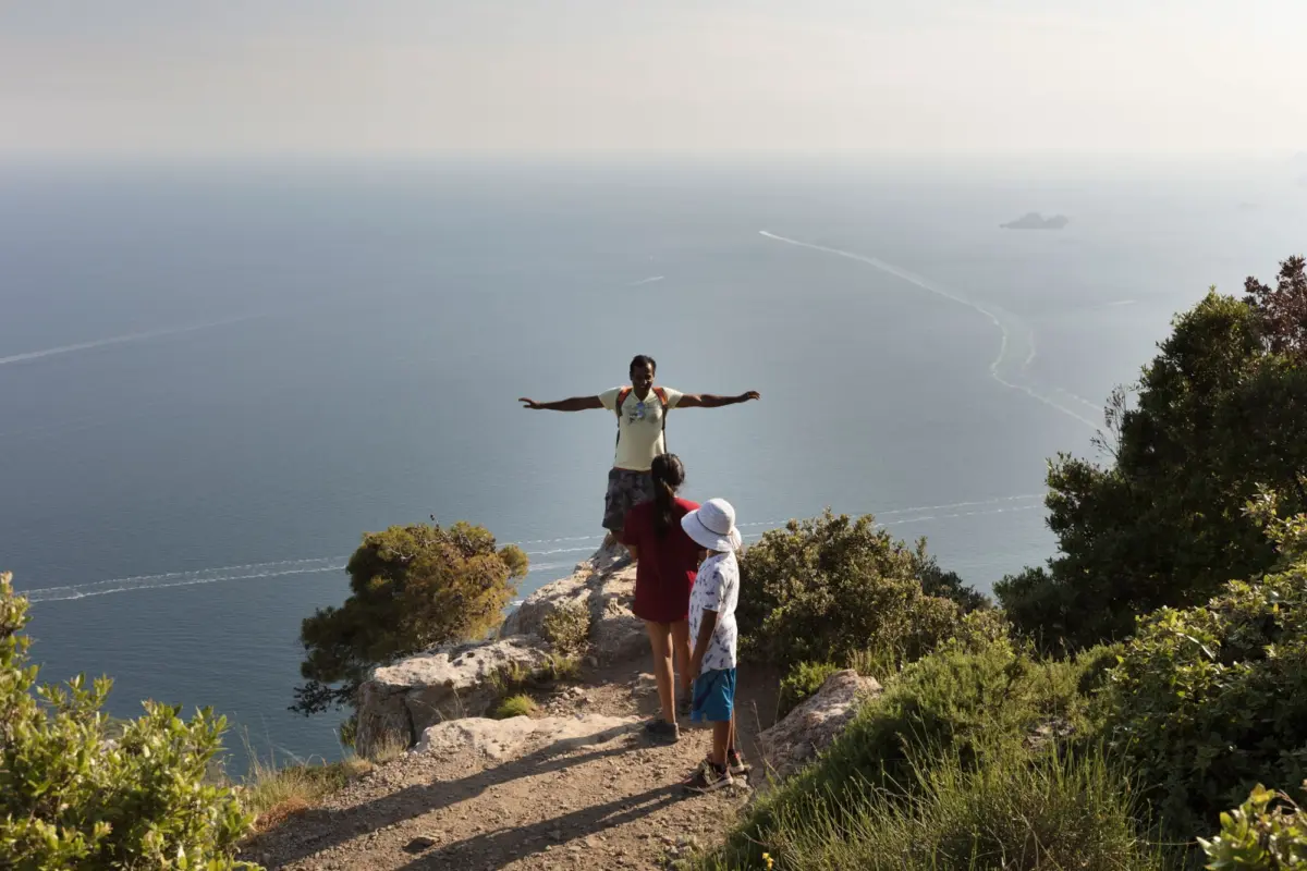 Man with arms outstretched on a rocky cliff above the sea; two children nearby, shrubs, distant boats on the horizon.
