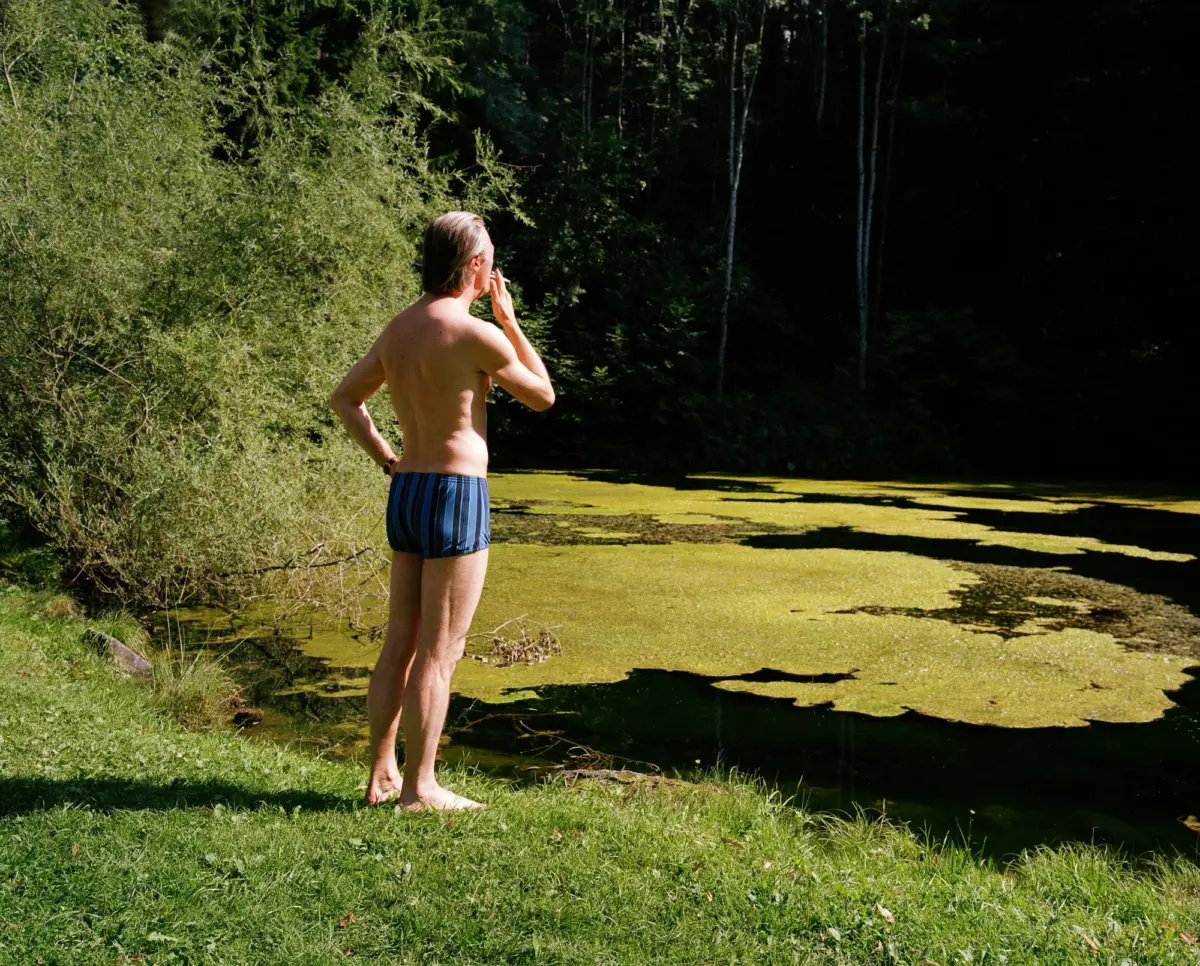 Adult person, shirtless, wearing striped swim trunks, stands beside an algae-covered pond at forest edge, hand to mouth.