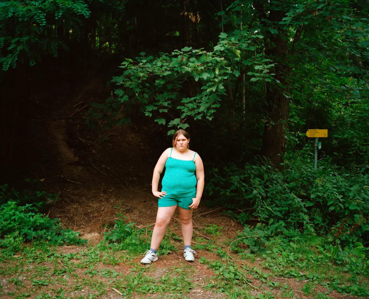 Young girl stands on a dirt forest trail, wearing a turquoise polka-dot tank top and matching shorts; dense greenery surrounds her.