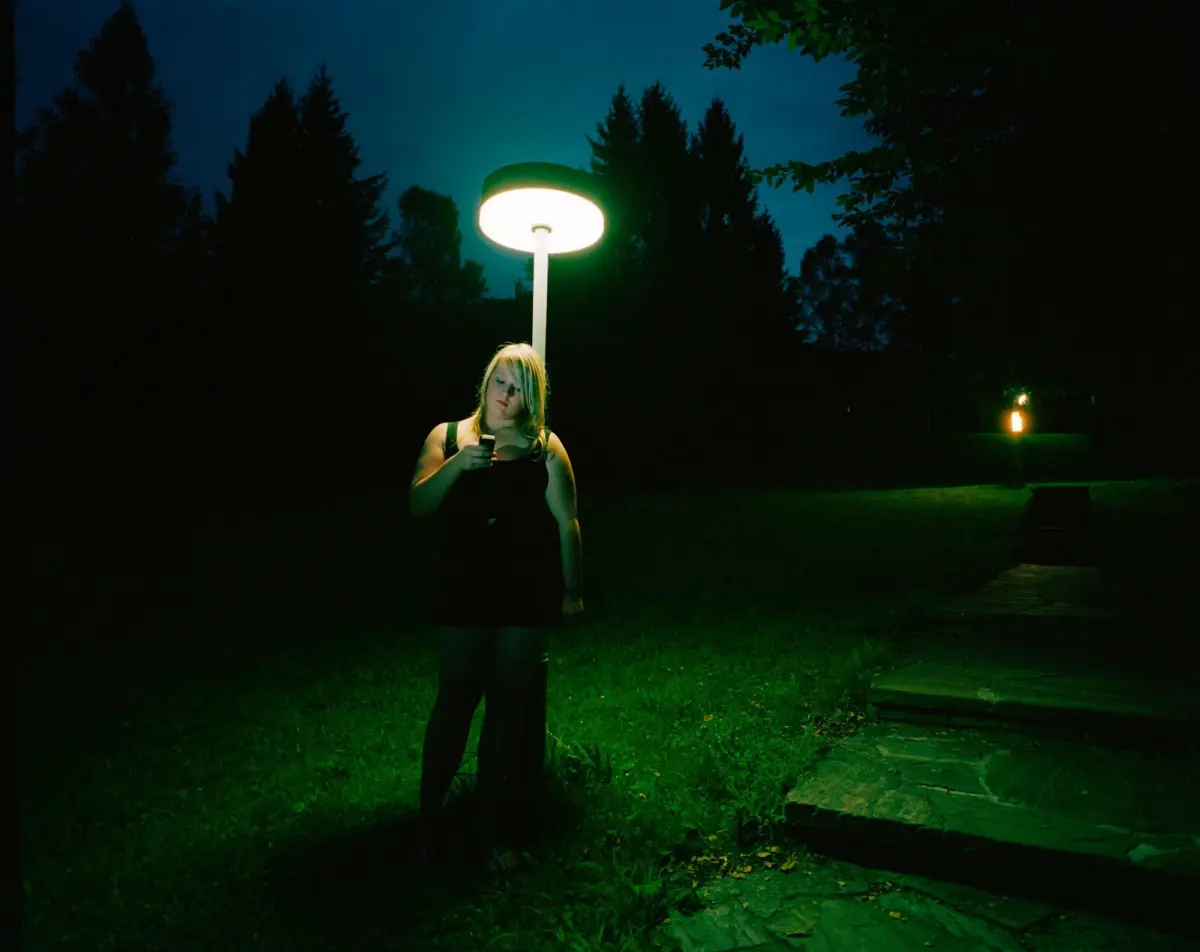 Person standing in a park at night beneath a glowing circular streetlamp, looking at a smartphone.