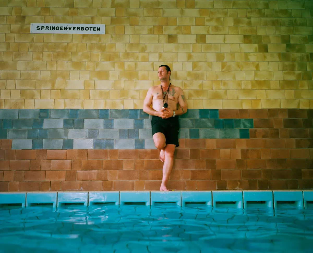 Shirtless tattooed man leaning against a tan tiled wall by a pool; sign reads SPRINGEN VERBOTEN.