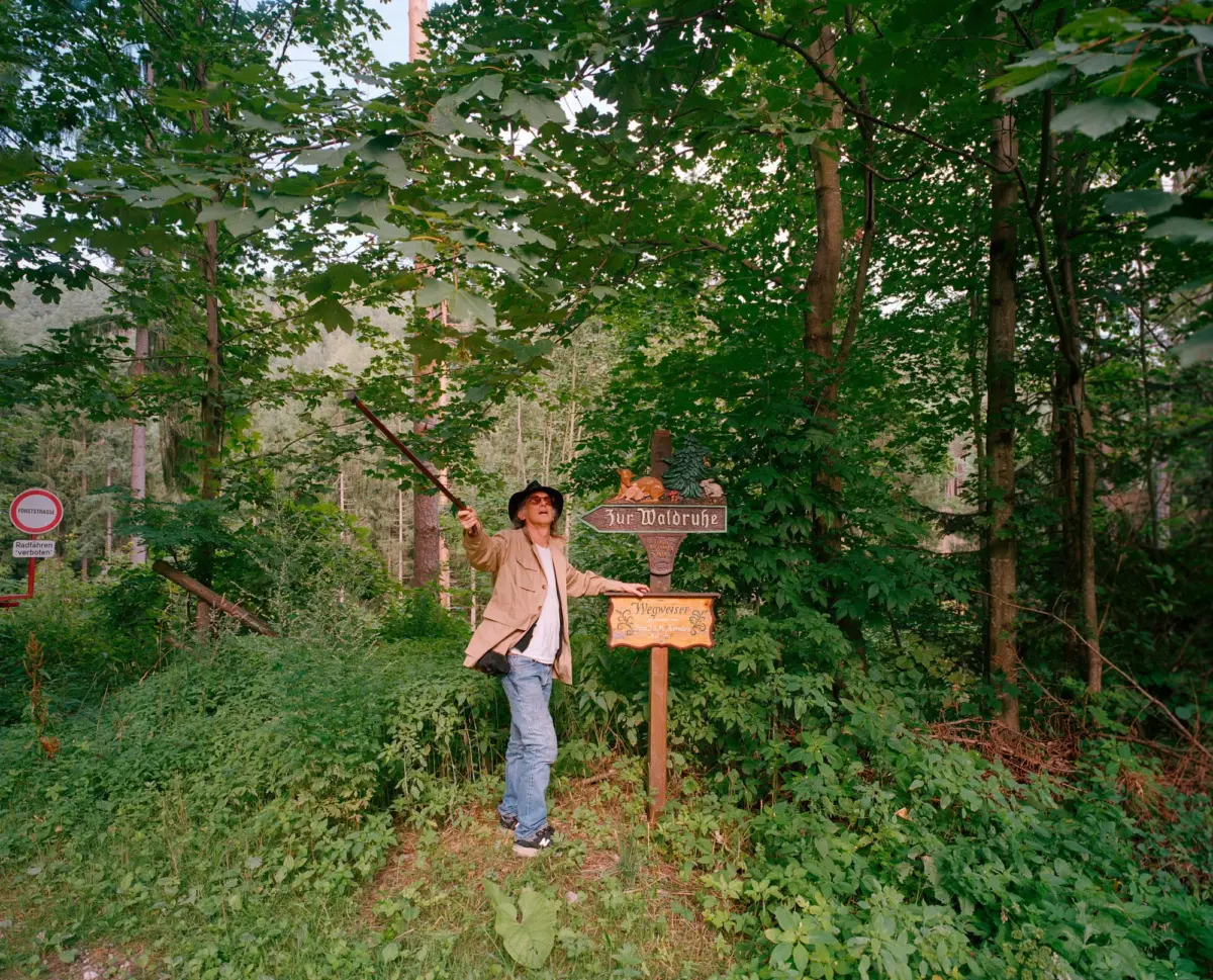 Adult man in a beige jacket and hat raises a long stick in a dense forest with wooden signposts.