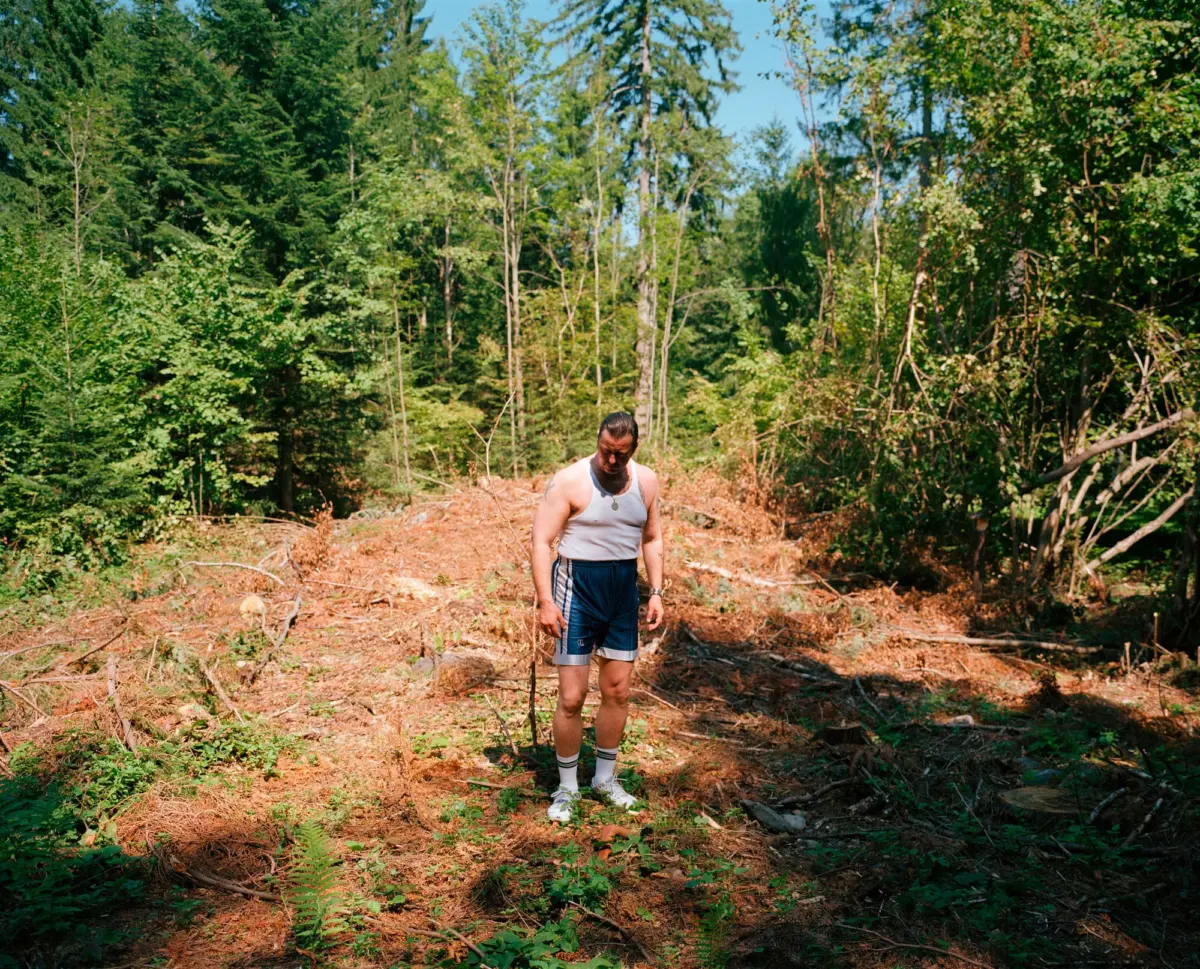 Adult person in a white sleeveless shirt and blue shorts stands in a sunlit forest clearing, looking down, holding a hiking stick.