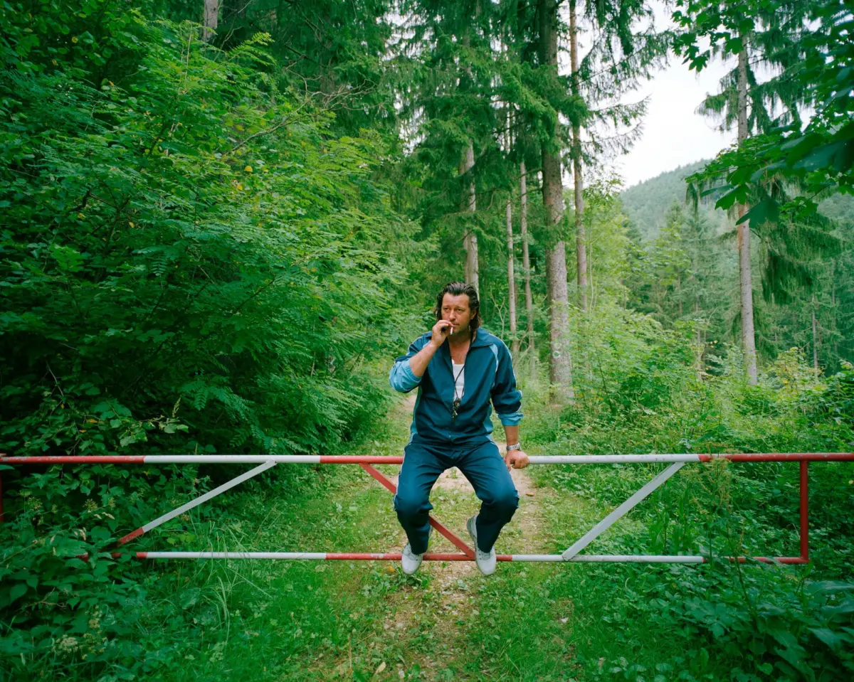 Person in blue tracksuit stands on a red-and-white barrier across a forest path, smoking.