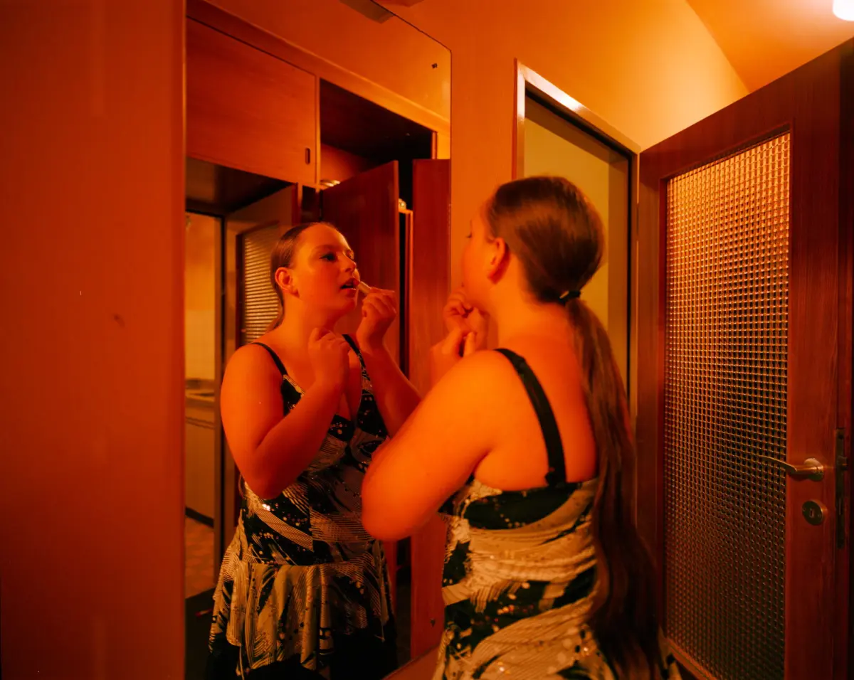 Two people in a warmly lit hallway, one applying lipstick before a mirror while the other watches.