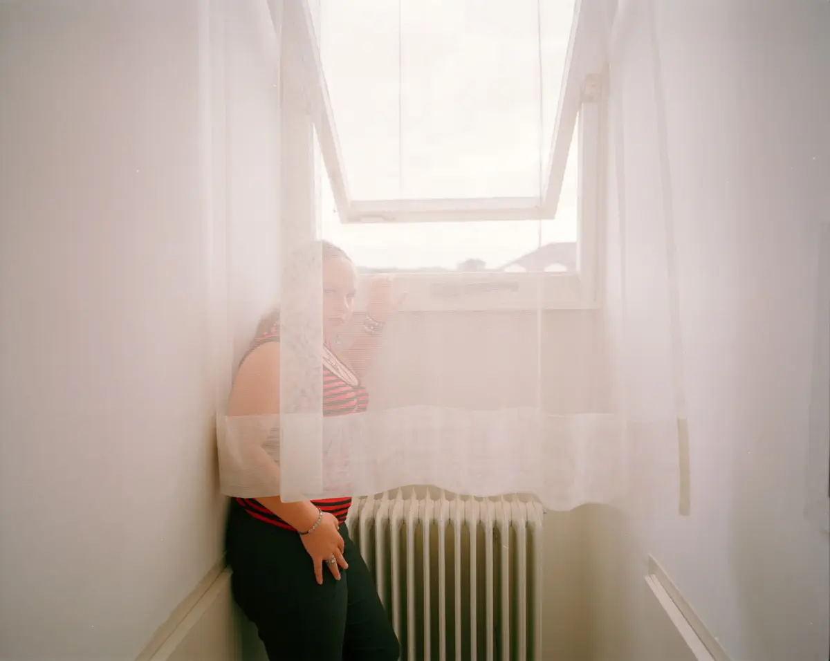Person in striped top and black pants stands in a narrow window alcove with an open window, sheer curtains, and radiator beneath.