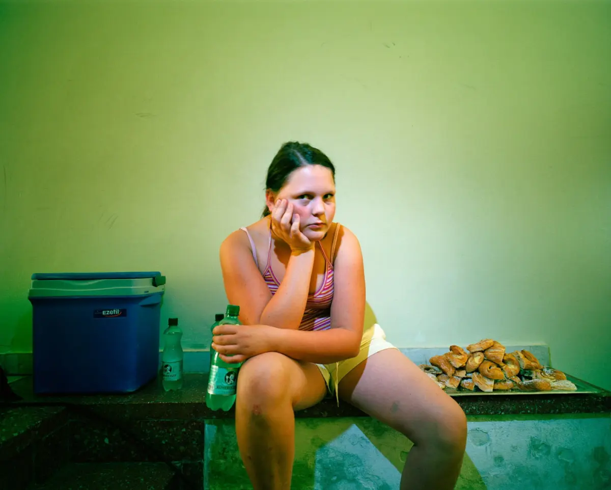 Person sits on steps by a pale green wall, resting chin on hand, holding a plastic bottle; blue cooler left, pile of bread rolls right.
