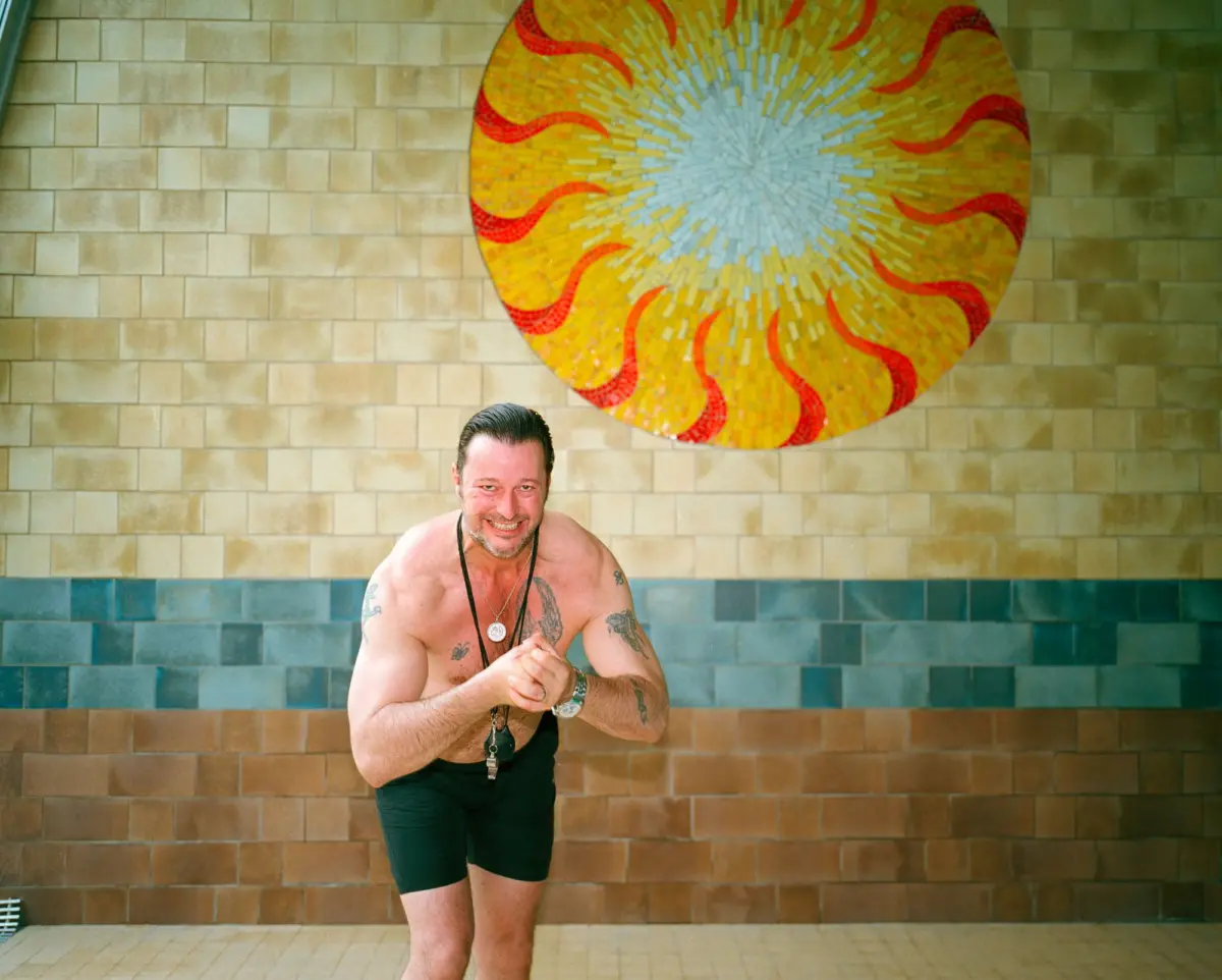 Smiling shirtless adult with tattoos and necklaces, standing with hands clasped in front of a beige-tiled wall and a large sun mosaic.