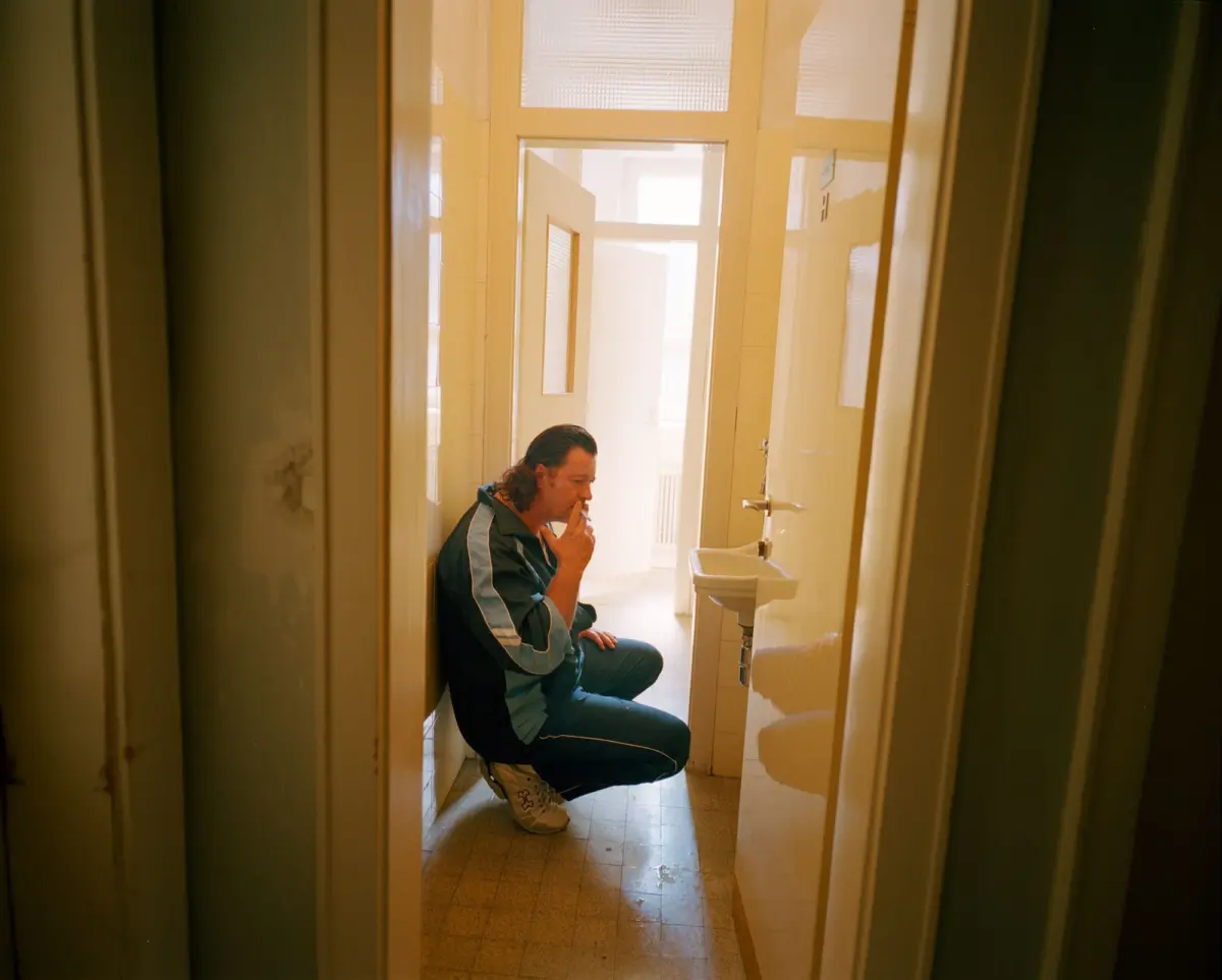 Person in teal tracksuit crouches in a narrow restroom, smoking a cigarette beside a sink.