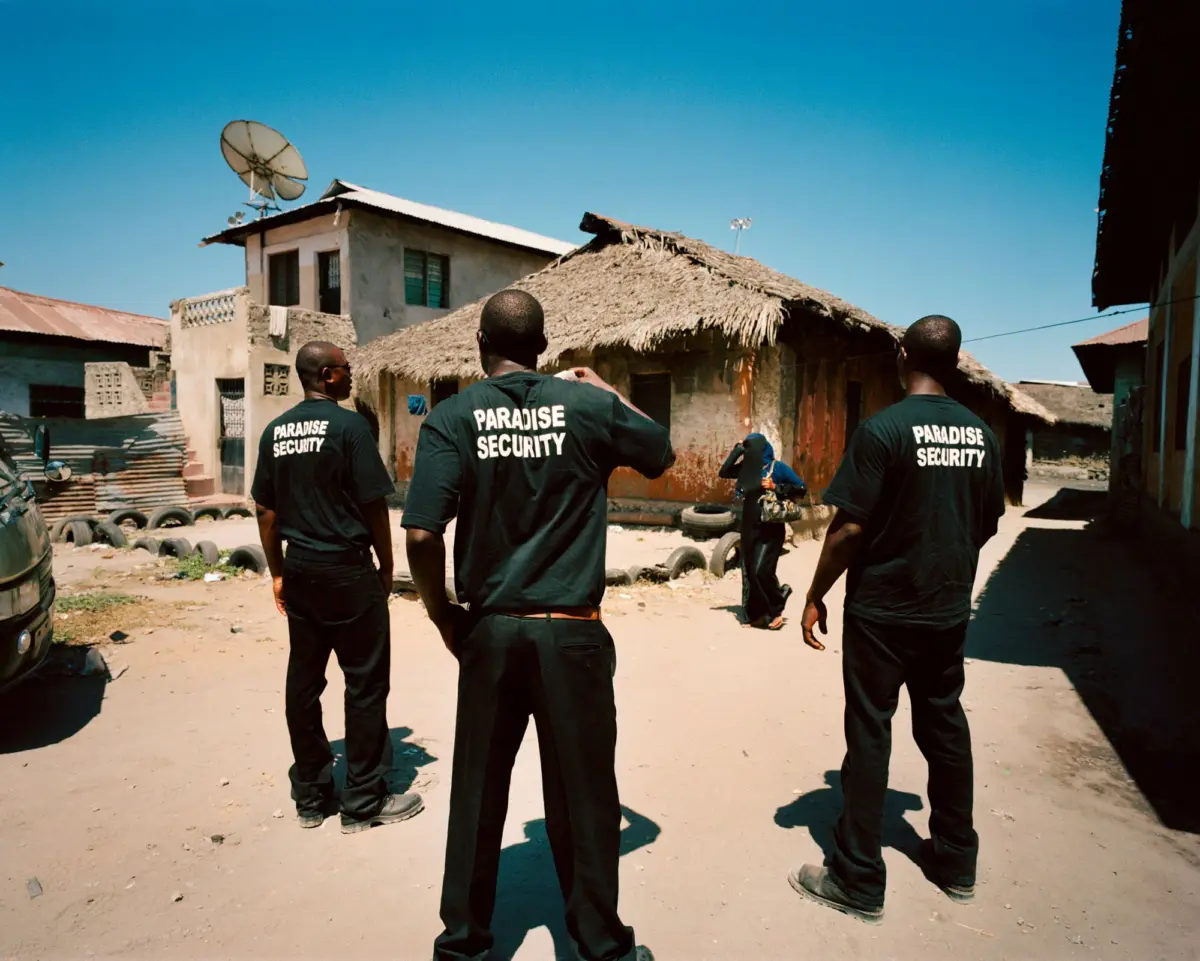 Three Paradise Security guards in black shirts stand with backs to the camera in a dusty street beside thatched huts.