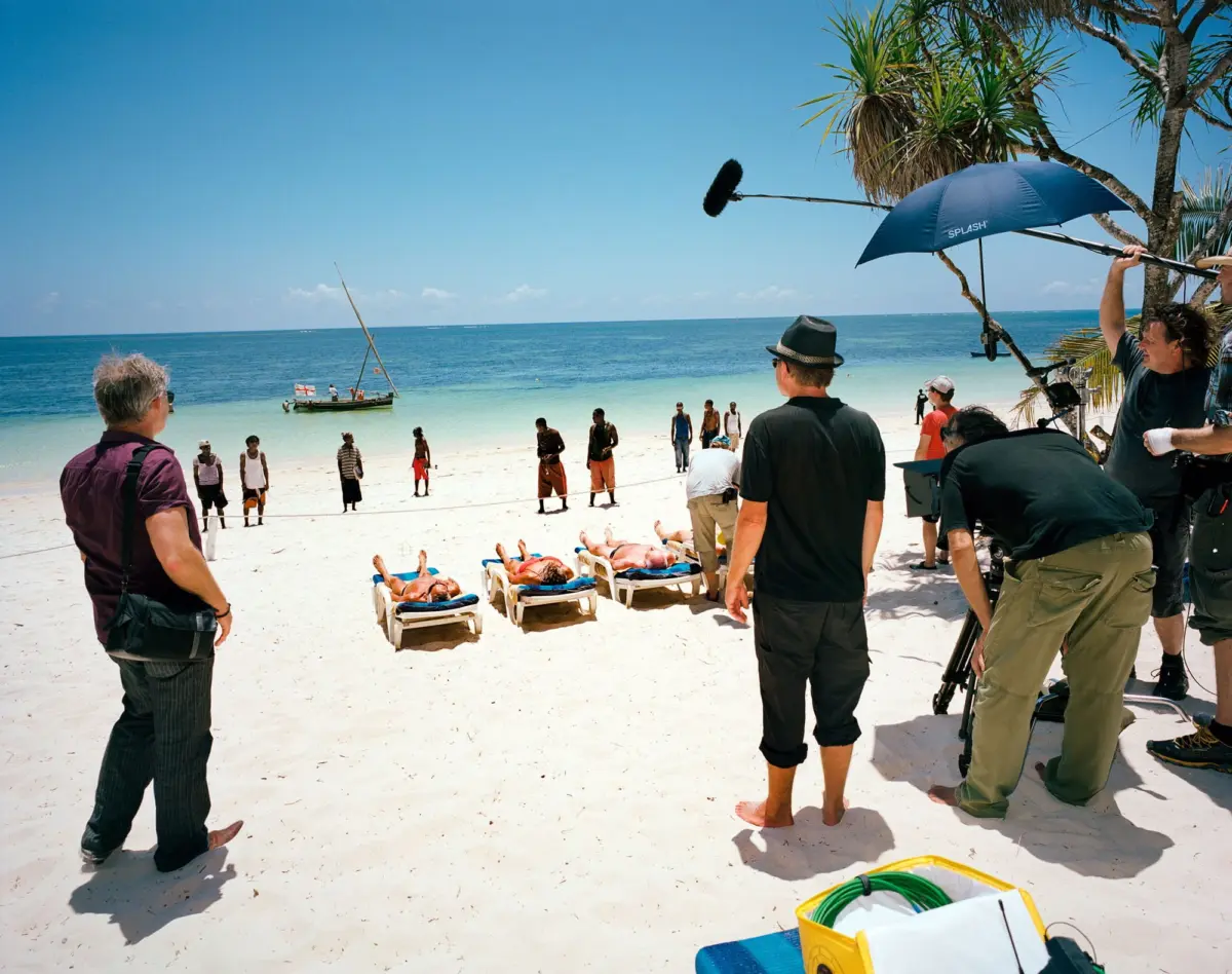 Film crew on sunny beach shoots ocean scene; boom microphone overhead, palm trees, sun loungers, sailboat in turquoise water.