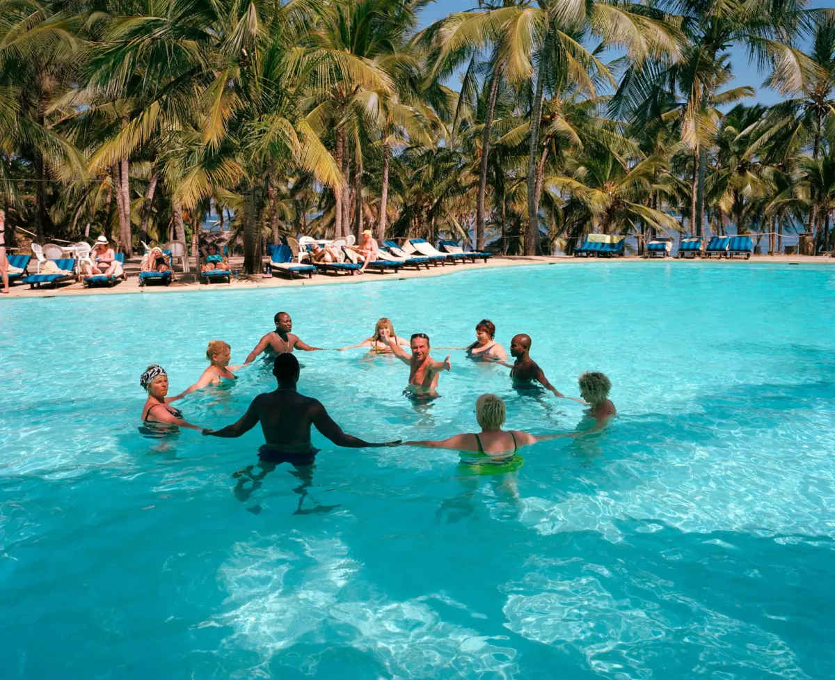 Group of adults swim and play in turquoise resort pool; palm trees and lounge chairs line the sunny poolside.