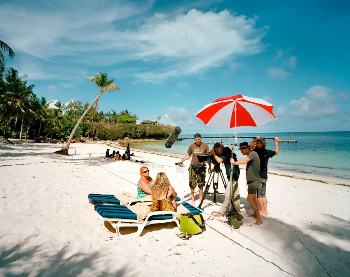 Film crew shoots on a tropical beach, setting up camera gear under a red-and-white umbrella.