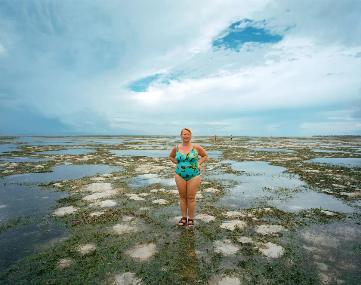 Adult person in turquoise patterned swimsuit stands on muddy flats at the beach under a cloudy sky.