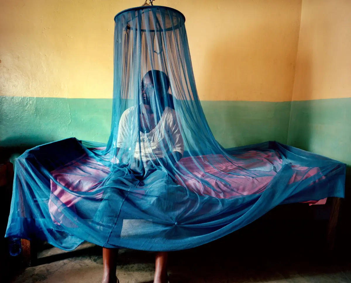 Person inside a blue mosquito net canopy indoors, standing by a bed with blue and pink sheets; yellow and green wall behind.
