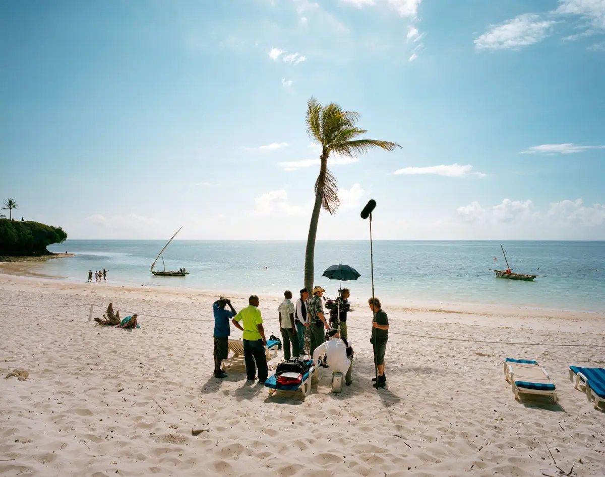 Film production crew setting up camera equipment on sunlit sandy beach near a palm tree and calm sea.