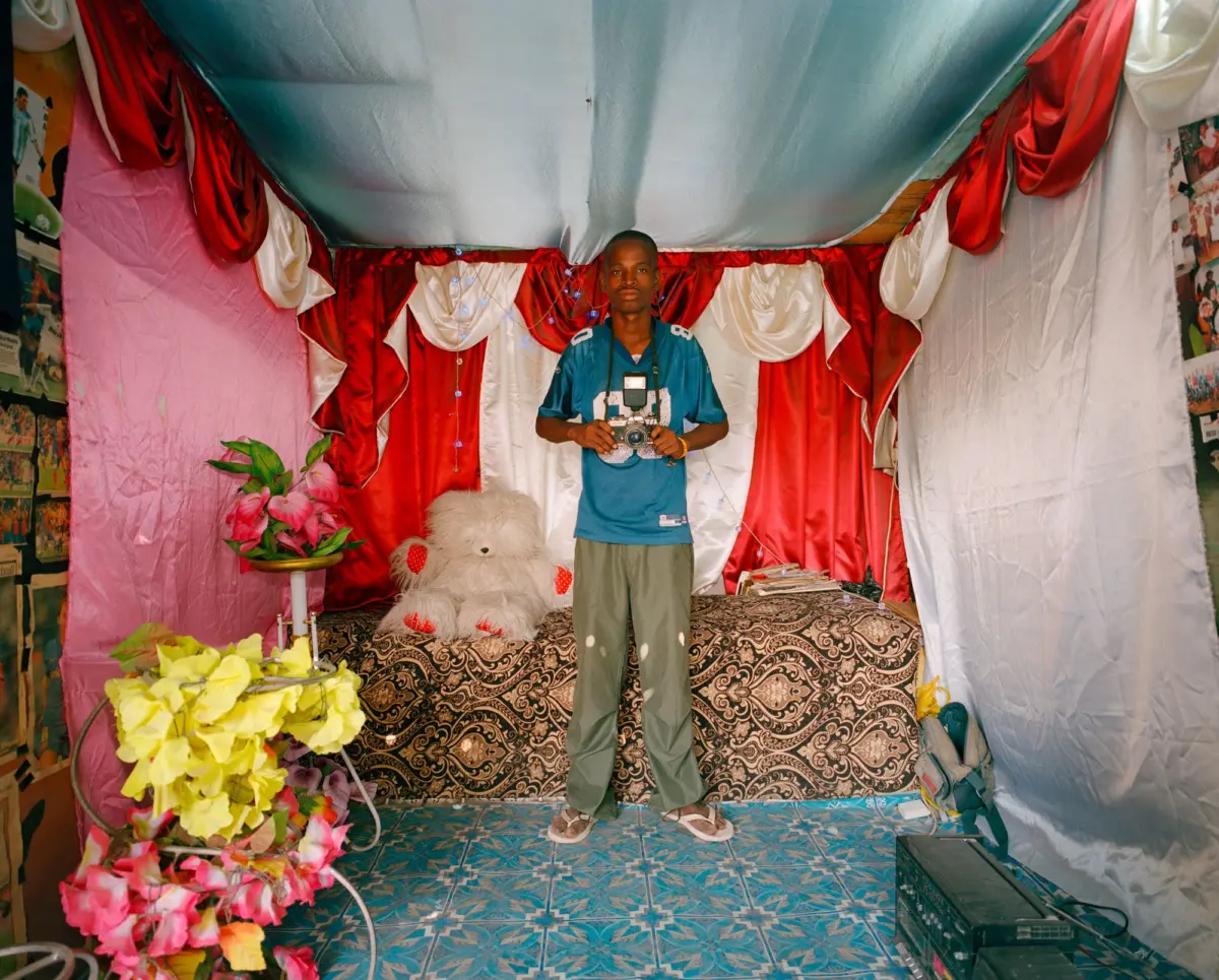 Person with camera stands in a festively decorated room with red and white drapes, pink wall, blue ceiling, and a large teddy bear on a patterned bed.