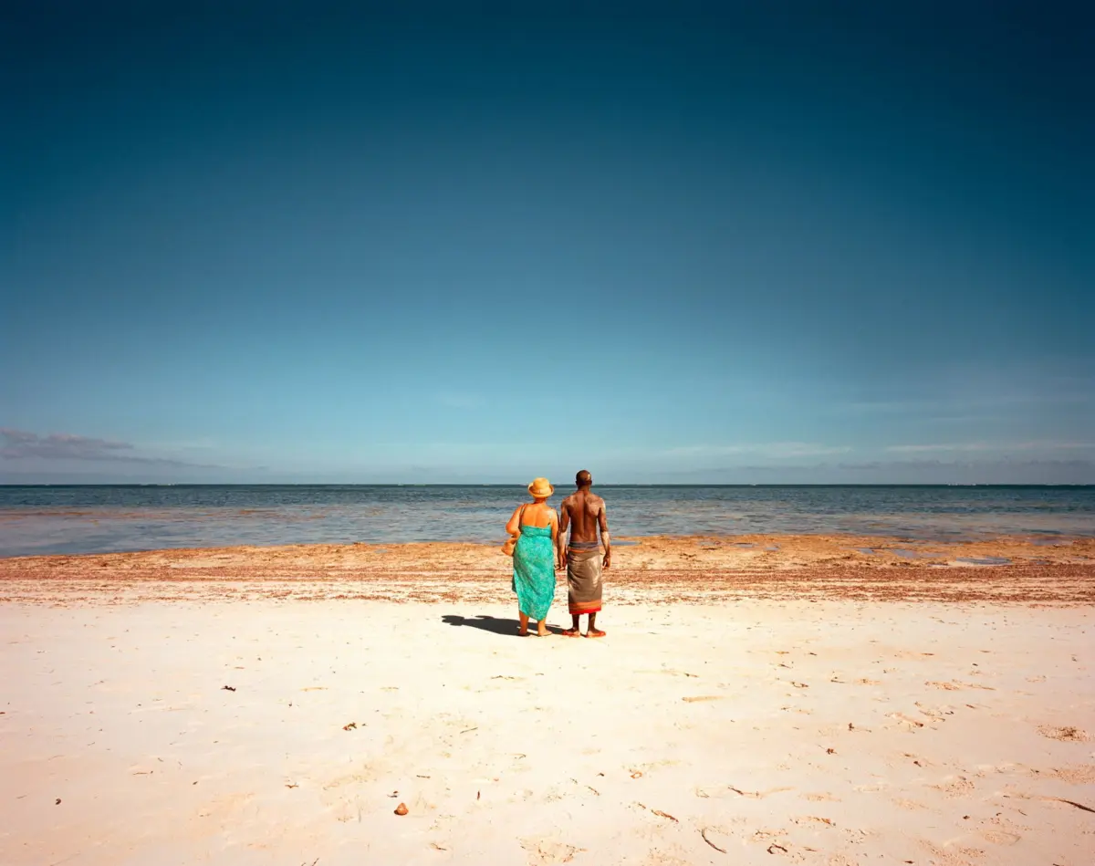 Two people stand at the water's edge on a sandy beach, facing the ocean under a clear blue sky.