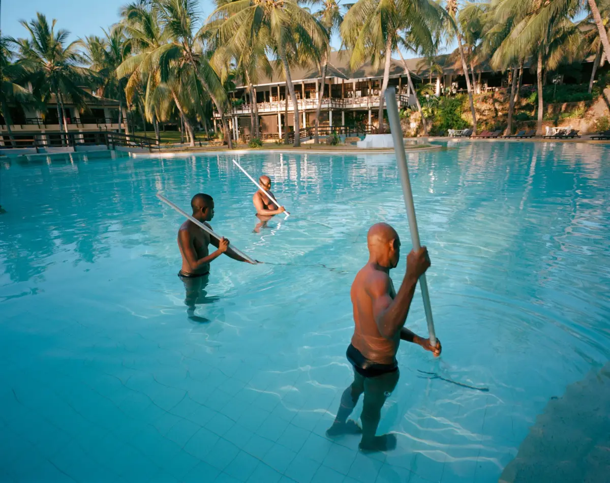 Three shirtless men waist-deep in a turquoise resort pool, holding long white poles, with palm trees and a resort building behind.