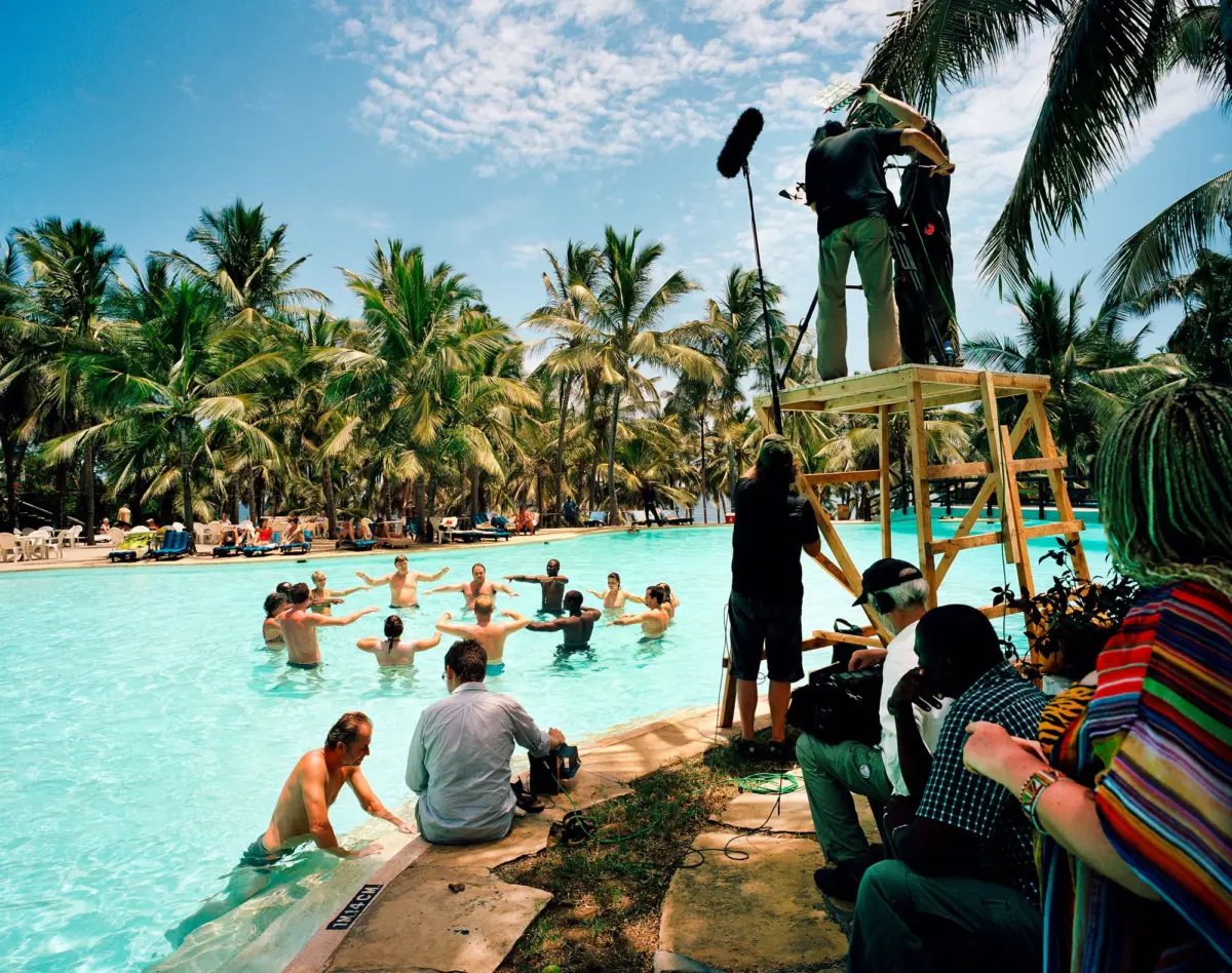 Film crew on a wooden platform films swimmers in a tropical pool, with a boom mic and palm trees under a blue sky.