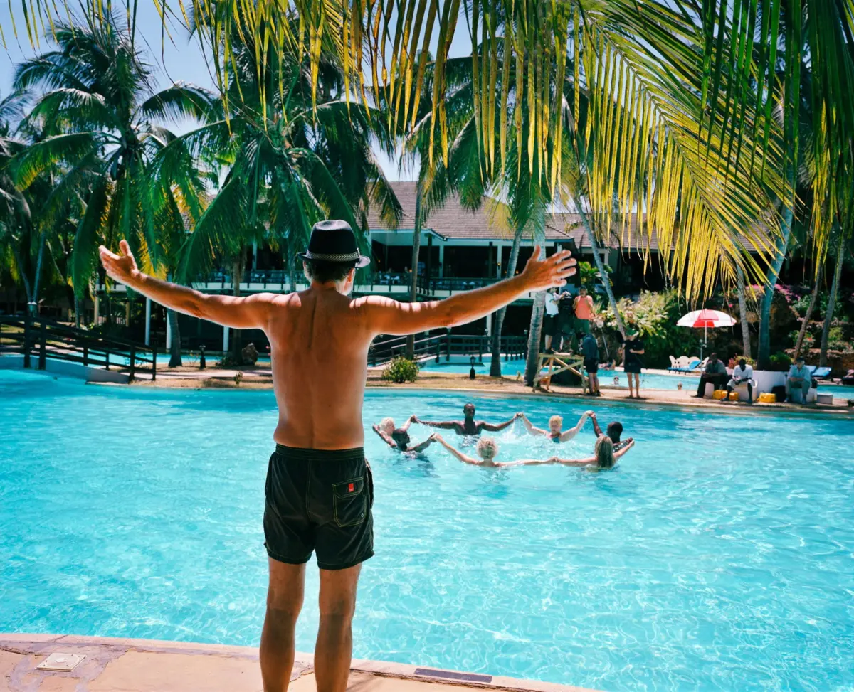 Back view of shirtless man in swim shorts wearing a hat, arms outstretched at pool edge, tropical palm trees and resort buildings in background.