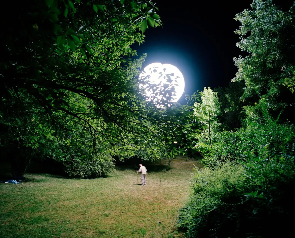 Bright full moon behind leafy trees in a park at night; a person stands on the grass.