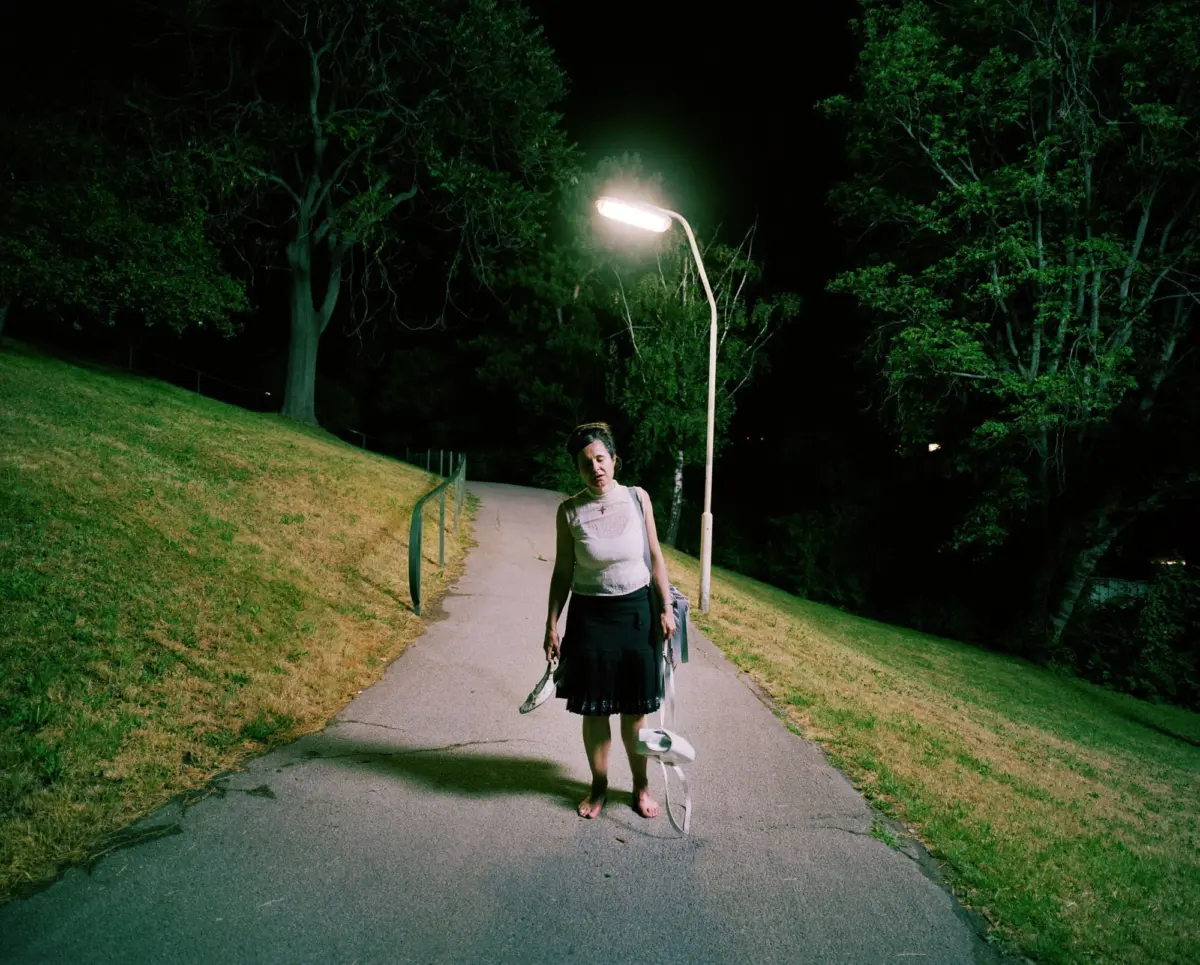 Night scene in a park; a person in a white sleeveless top and black skirt stands barefoot on a path beside a glowing streetlamp.