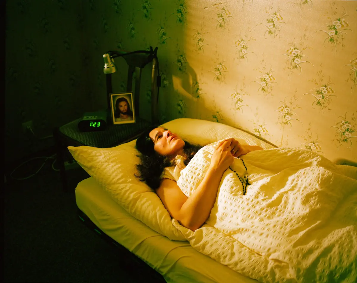 Bearded person lying in bed under a quilt in a bedroom with floral wallpaper; hands clasped, beside a digital alarm clock and Jesus portrait.