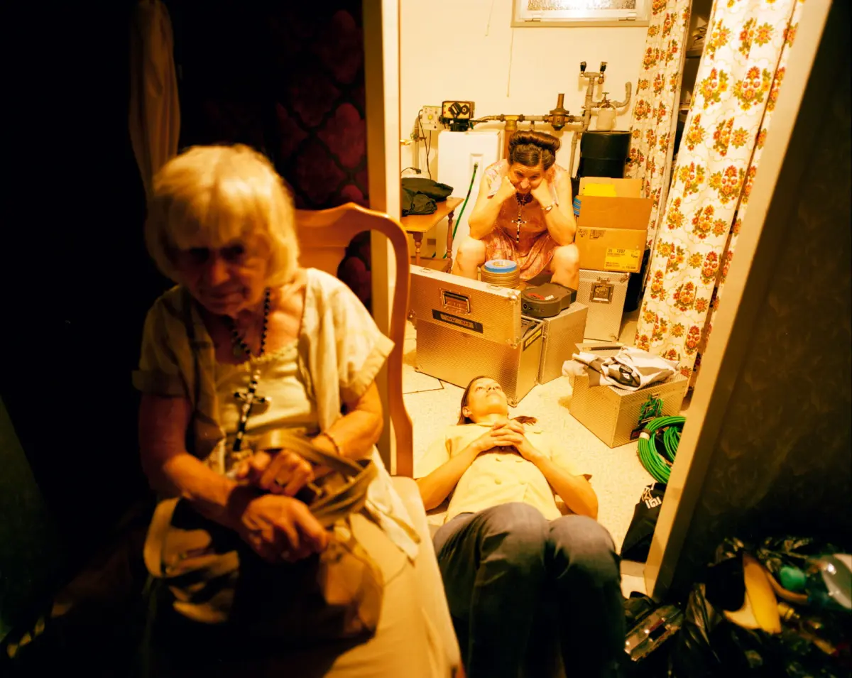 Person in pink dress sits distressed on cluttered storage-room floor with hands on head; others rest among boxes, pipes, and floral curtains.