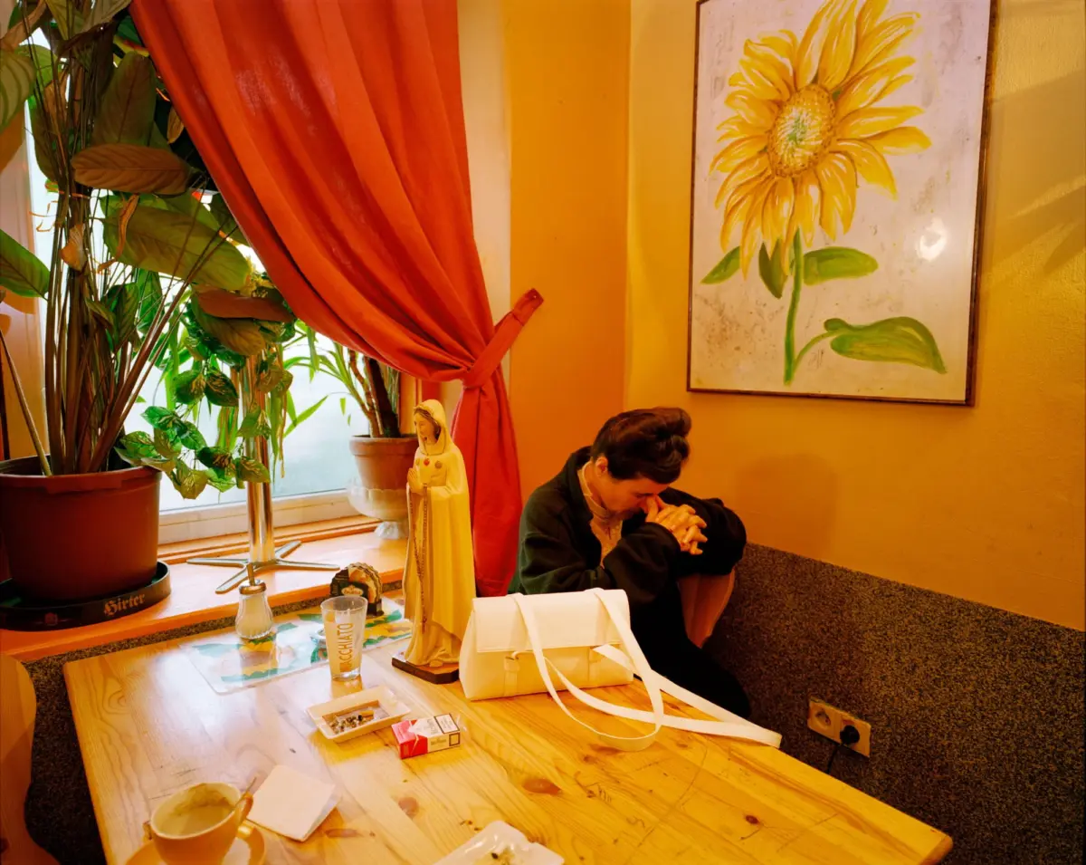 Two people sit at a wooden table in a warm cafe; one leans forward with head down near a sunflower painting, orange walls, and potted plants.