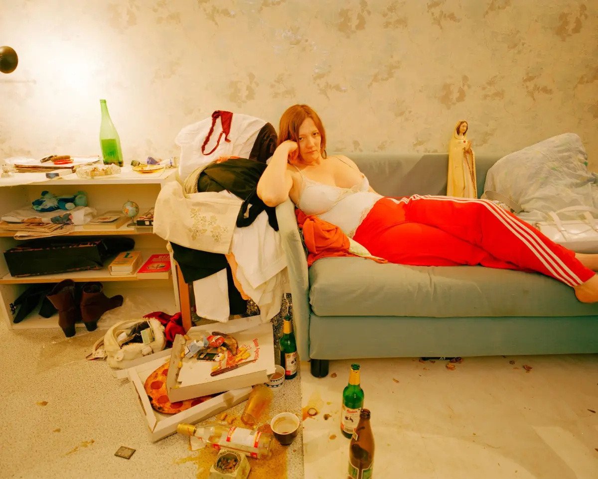 Red-haired person lounging on a messy bed in a cluttered room with peeling wallpaper, bottles and papers scattered.
