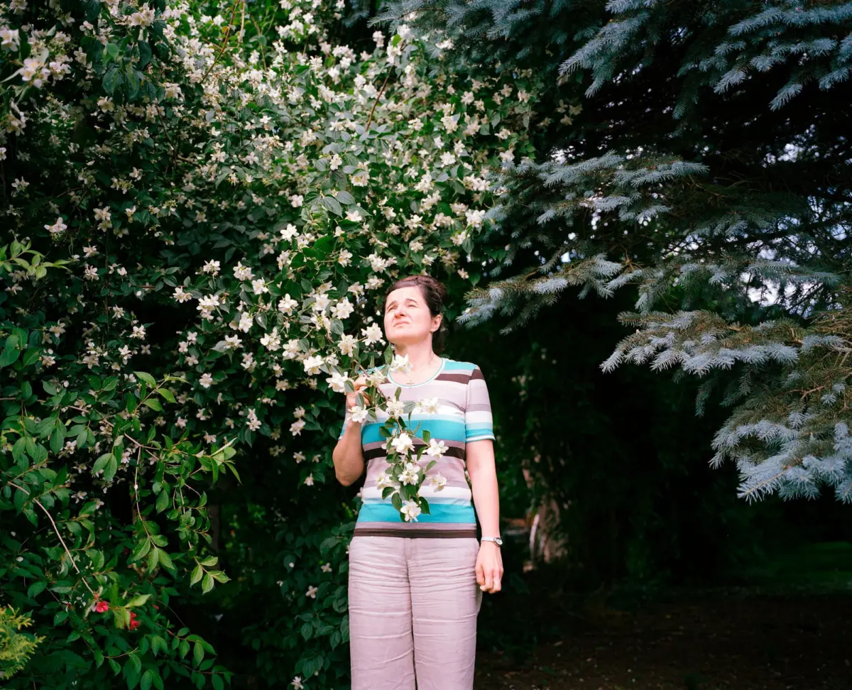 Adult woman standing among a white-flowering shrub and blue spruce in a garden, looking upward.